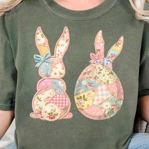 Könnte beinhalten: Grünes T-Shirt mit zwei Patchwork-Hasen-Designs. Ein Hase steht aufrecht, der andere ist eiförmig. Beide Hasen haben Blumen- und Vichy-Muster in Rosa, Blau und Gelb. Das Shirt hat eine lockere Passform.