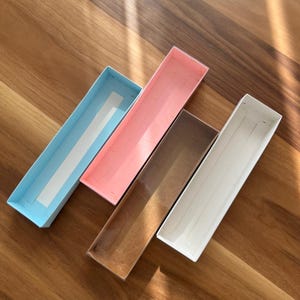 Puede incluir: Cuatro cajas rectangulares abiertas en colores pastel: azul claro, rosa, marrón y blanco. Están dispuestas en diagonal sobre una superficie de madera. La caja azul claro tiene una franja blanca. Las cajas están vacías.