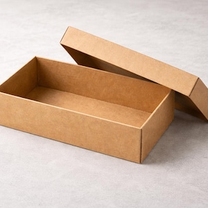 Puede incluir: Una caja de cartón rectangular abierta, con la tapa ligeramente entreabierta. La caja y la tapa son de color marrón claro, y la caja está vacía. La caja está sobre una superficie gris claro.
