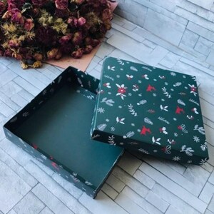 Può includere: Una scatola regalo quadrata aperta, con interno ed esterno verde scuro. Il coperchio presenta un motivo ripetuto di foglie bianche, bacche rosse e fiocchi di neve su sfondo verde scuro. La scatola è vuota.