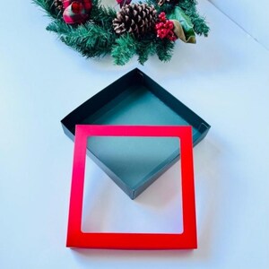May include: An open black gift box with a red frame and a green wreath with pinecones and red berries in the background. The box has a dark teal interior.