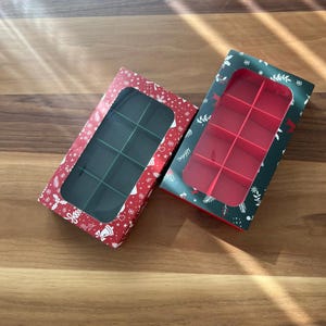 Könnte beinhalten: Zwei weihnachtliche Geschenkboxen. Eine rote Box mit weißen Schneeflocken und grünem Innenraum, die andere grün mit weißen Blumenmustern und rotem Innenraum. Beide haben transparente Fenster und Fächer.