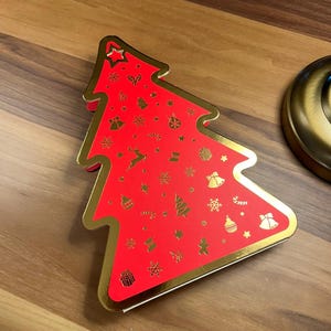 May include: A red Christmas tree-shaped box with gold trim and festive gold icons. The box is decorated with stars, bells, snowflakes, and other holiday-themed designs. The box is on a wooden surface.