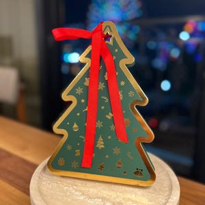 May include: A green Christmas tree-shaped gift box with gold trim and a red ribbon. The box is decorated with festive holiday icons and sits on a round, light-colored stone tray. The background is blurred with city lights.