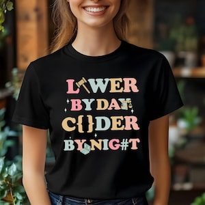 Könnte beinhalten: Schwarzes T-Shirt mit dem Text "Lawyer by Day, Coder by Night" in Pastellfarben. Das Design beinhaltet einen Hammer, eine Waage und ein Laptop-Symbol. Das T-Shirt hat einen Rundhalsausschnitt.