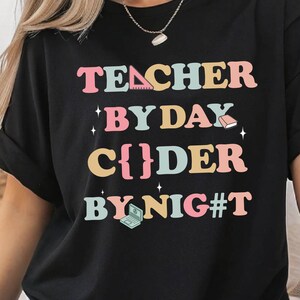 Könnte beinhalten: Schwarzes T-Shirt mit dem Text "TEACHER BY DAY C{}DER BY NIG#T" in bunten Buchstaben. Das Design beinhaltet Symbole für Lineal, Buch und Laptop. Das Shirt hat einen lässigen Stil.