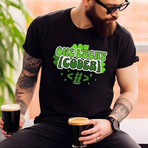 Könnte beinhalten: Schwarzes T-Shirt mit dem Text "ONE LUCKY {CODER}" in Grün und Weiß. Das Design beinhaltet ein Kleeblatt und Codesymbole. Die Person im Bild hält zwei Gläser dunkles Bier.