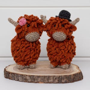 Puede incluir: Dos figuras de crochet hechas a mano de vacas de las Tierras Altas, una con una flor rosa y la otra con un sombrero de copa negro, están sobre una rodaja de madera. Las vacas son marrones con cuernos y caras beige.