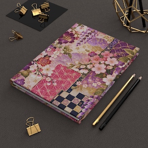Pode incluir: Um caderno retangular com um design de patchwork vibrante de flores de cerejeira cor-de-rosa, glicínias roxas e padrões geométricos em tons de magenta, azul-marinho e dourado. A capa exibe uma mistura de motivos florais e formas tradicionais.