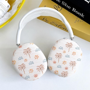 May include: Over-ear headphones featuring a white headband and ear cups decorated with a repeating pattern of brown palm trees, orange hibiscus flowers, blue wavy lines, and small seashells against a light cream-colored background.