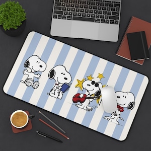 May include: A rectangular desk mat featuring a light blue and white vertical striped pattern with four illustrations of Snoopy. The character is shown sitting, reading a book, playing a red guitar with yellow stars, and holding a red flower.