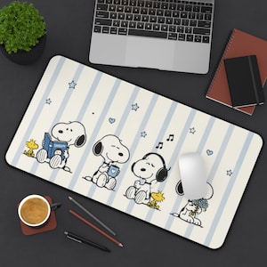Può includere: Un tappetino da scrivania rettangolare con uno sfondo color crema chiaro e strisce verticali azzurre. Il tappetino mostra quattro illustrazioni di Snoopy in varie attività, tra cui leggere un libro con l'etichetta Snoopy, tenere una tazza, indossare cuffie e tenere un bouquet di fiori blu, accompagnato da Woodstock e piccole stelle blu.