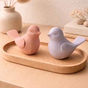 Op de afbeelding: Twee keramische vogelbeeldjes in roze en lavendel op een houten dienblad. De vogels hebben afgeronde lichamen, kleine snavels en gedetailleerde vleugels. De lade is lichtbruin en de achtergrond bevat een vaas met gedroogde bloemen.