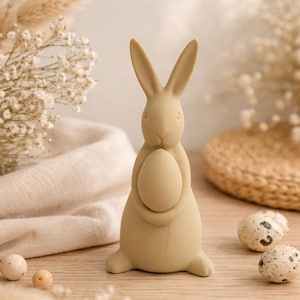 Puede incluir: Una figura de conejo beige sosteniendo un huevo, un artículo decorativo para la Pascua. El conejo tiene orejas largas y un acabado liso. En la imagen también hay pequeños huevos moteados y flores secas.