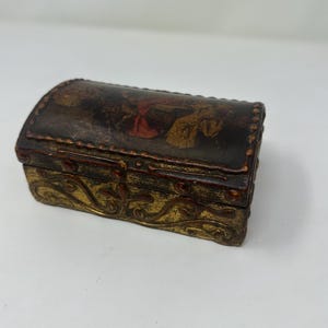 May include: A small, antique-style box with a curved lid. The box is primarily dark brown with gold accents and a decorative design. The lid features a faded painting with red and gold hues. The box is likely made of wood or a similar material.