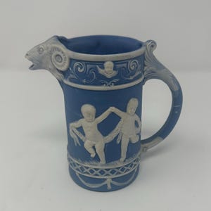 May include: Blue ceramic pitcher with a ram's head spout and a decorative handle. The pitcher features white relief figures of cherubs and ornate detailing. A classic piece for serving beverages.
