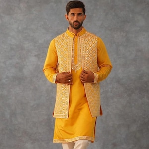 Pode incluir: Um homem veste um kurta amarelo mostarda e um colete bordado a condizer sobre calças creme. O traje apresenta uma gola mandarim e acabamentos decorativos. Os sapatos são de cor bege claro. O fundo é cinzento liso.