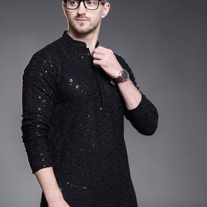 Könnte beinhalten: Ein schwarzer, langärmeliger Kurta mit Stehkragen und aufwendiger Ton-in-Ton-Stickerei auf dem gesamten Stoff. Das Kleidungsstück ist mit kleinen, verstreuten Pailletten verziert, die das Licht einfangen und einen subtilen Schimmer erzeugen.