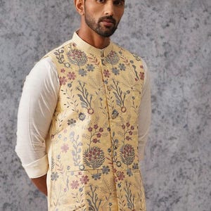 May include: A sleeveless vest in a pale yellow hue featuring a detailed floral pattern in shades of blue, red, and grey. The garment has a mandarin collar, a front button closure with metallic buttons, and two lower pockets on the front panel.