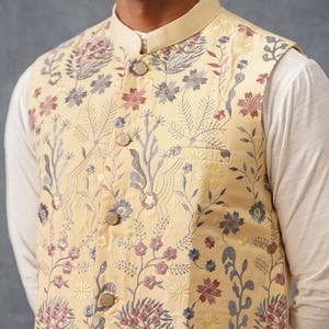 May include: A pale yellow sleeveless vest featuring a mandarin collar and a front button closure. The garment is decorated with an intricate floral and botanical embroidery pattern in shades of muted pink, grey, and yellow, with a chest pocket.