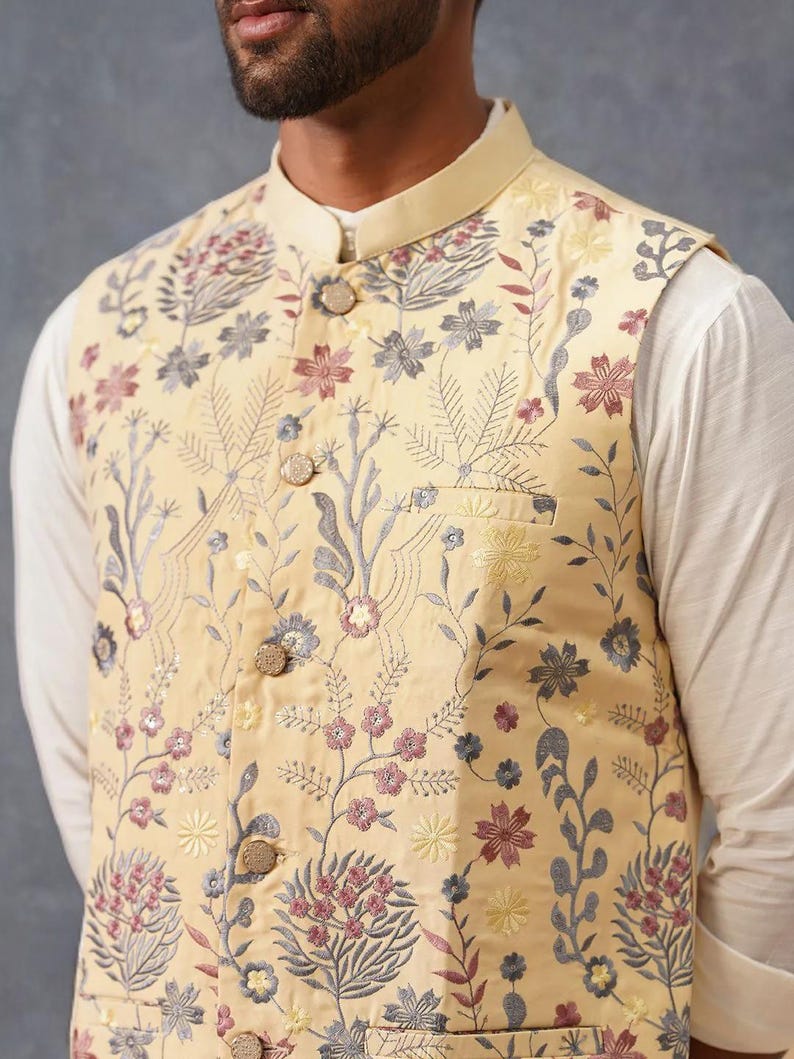 May include: A pale yellow sleeveless vest featuring a mandarin collar and a front button closure. The garment is decorated with an intricate floral and botanical embroidery pattern in shades of muted pink, grey, and yellow, with a chest pocket.