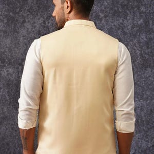 May include: A sleeveless beige vest worn over a long-sleeved white shirt. The vest features a high collar and a smooth, solid-colored fabric finish. The white shirt has sleeves rolled up to the elbows, and the ensemble is viewed from the back.