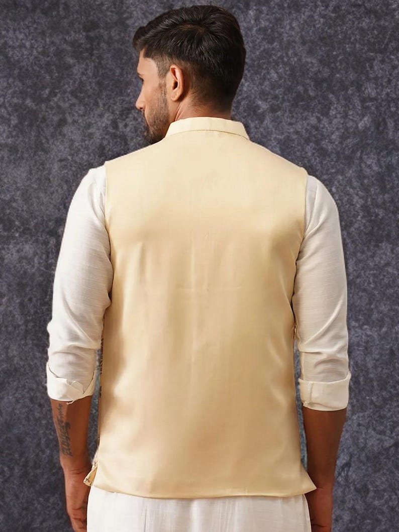 May include: A sleeveless beige vest worn over a long-sleeved white shirt. The vest features a high collar and a smooth, solid-colored fabric finish. The white shirt has sleeves rolled up to the elbows, and the ensemble is viewed from the back.