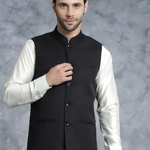 May include: A black Nehru-collared waistcoat with four buttons and two pockets, worn over a white long-sleeved shirt. The waistcoat is tailored and appears to be made of a smooth fabric. The background is a mottled grey.