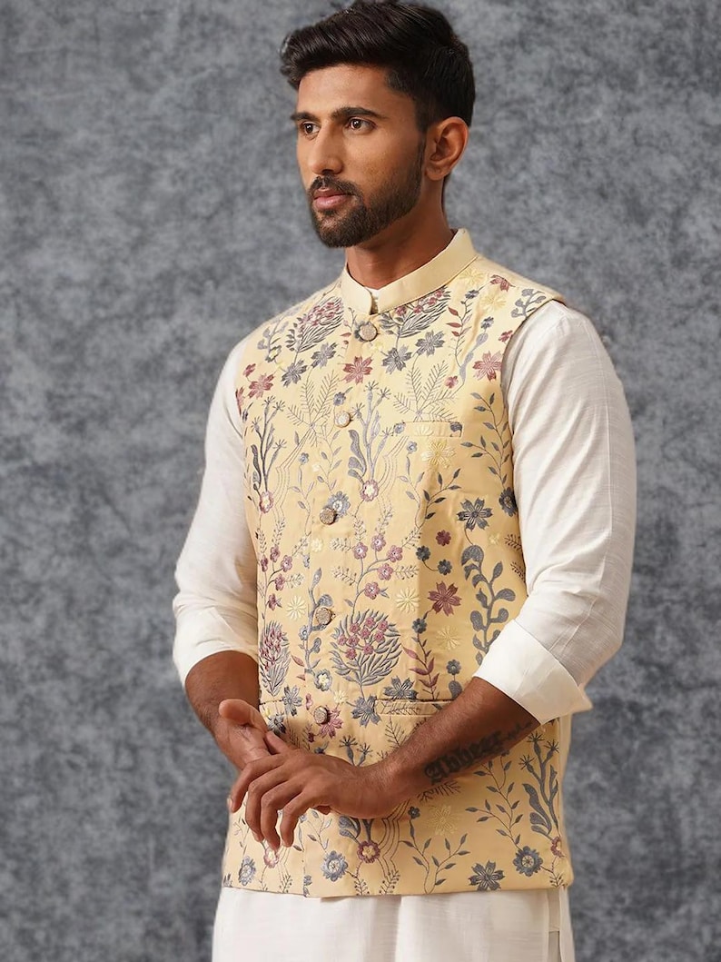 May include: A light yellow sleeveless vest featuring a mandarin collar and a button-down front. The garment is decorated with an intricate floral pattern in shades of blue, pink, and grey. It is worn over a plain white long-sleeved shirt.