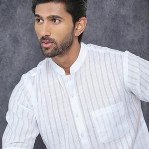 May include: A white, striped shirt with a mandarin collar and a single chest pocket. The shirt has a button-down placket and long sleeves. The fabric appears to be lightweight and breathable, suitable for warm weather.