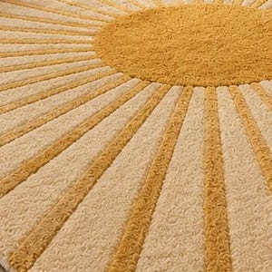 May include: A circular area rug featuring a textured, tufted surface with a central mustard yellow circle and radiating sunburst rays in alternating mustard yellow and cream white colors, creating a geometric pattern across the entire rug.
