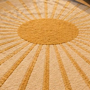 May include: A circular rug featuring a central mustard yellow circle with a textured surface, surrounded by alternating radiating rays of mustard yellow and cream white. The entire design has a consistent, dense, and tufted pile texture throughout.