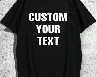 May include: Black t-shirt with the words "CUSTOM YOUR TEXT" in bold white letters. The shirt is hanging on a wooden hanger against a textured gray background. The short-sleeved tee has a crew neck.