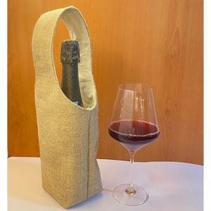 May include: A beige burlap wine bottle tote with a handle, holding a dark bottle. A clear wine glass with red wine sits next to the tote. The background is a light brown wooden surface.