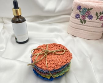 May include: A stack of colorful, crocheted facial rounds tied with twine. The rounds are in shades of orange, purple, green, blue, and yellow. A brown glass bottle with a dropper and a floral-patterned pink towel are in the background.