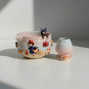 May include: A ceramic mixing bowl and a small matching cup featuring a pastel gradient of blue and pink. The bowl displays a raised figure of a girl flying on a broom with a black cat, a red radio, and bread loaves. The cup has paw prints and bread.