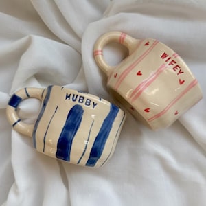 Puede incluir: Dos tazas de cerámica. Una es de color crema con rayas verticales azules y la palabra "Hubby". La otra es de color crema con rayas rosas, pequeños corazones rojos y la palabra "Wifey". Ambas tazas tienen asas redondeadas.