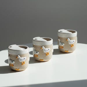 May include: Three ceramic travel mugs with a beige speckled finish and white lids. Each mug is adorned with raised white and yellow duck figures. The mugs are cylindrical and sit on a white surface.