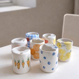 May include: A collection of seven white ceramic toothbrush holders. Each holder is decorated with a different fruit or vegetable design, including carrots, oranges, lemons, and blueberries. The holders are cylindrical with a curved top.