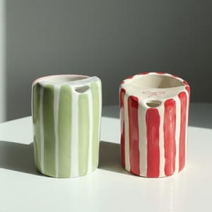 Può includere: Due contenitori in ceramica a righe verticali. Uno è verde a righe bianche, l'altro è rosso a righe bianche. Entrambi hanno una piccola apertura nella parte superiore e un piccolo foro laterale.