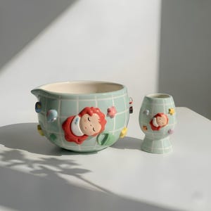May include: A pair of ceramic planters in a light green grid pattern. The larger, bowl-shaped planter has a spout. Both planters feature a sleeping child design and colourful embellishments. The smaller planter is vase-shaped.