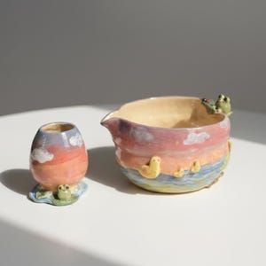 Op de afbeelding: Een keramische creamer en kleine vaas set. De creamer heeft een kikker- en eendjesontwerp. De vaas heeft een zonsondergang- en wolkenontwerp. De set is met de hand beschilderd met pastelkleuren.