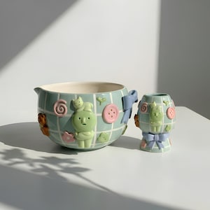 Puede incluir: Un juego de cuenco y jarrón de cerámica caprichoso. Las piezas de color verde claro presentan un patrón de cuadrícula, con adornos 3D de personajes de dibujos animados, botones, lazos y flores. El cuenco tiene un pico y un asa. El jarrón tiene una forma más pequeña y redondeada.