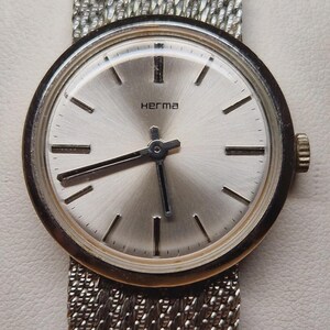 Puede incluir: Un reloj de pulsera vintage con esfera y agujas plateadas, con la marca "Herma". El reloj tiene una correa de malla metálica y un bisel dorado. La esfera del reloj tiene marcadores de hora sencillos y elegantes.
