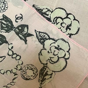 May include: Two light pink handkerchiefs with black and white floral and bow designs. One handkerchief has a pink stitched border. The design includes a bow, a necklace, and a flower.