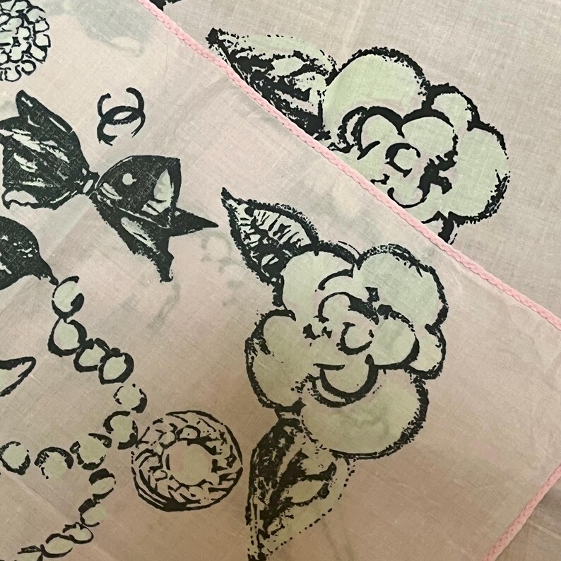May include: Two light pink handkerchiefs with black and white floral and bow designs. One handkerchief has a pink stitched border. The design includes a bow, a necklace, and a flower.