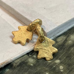 Op de afbeelding: Een paar goudkleurige oorbellen met een getextureerde, organisch gevormde bladhanger die aan een dikke, ronde ring hangt. Het metalen oppervlak heeft een matte, gehamerde afwerking met onregelmatige randen.