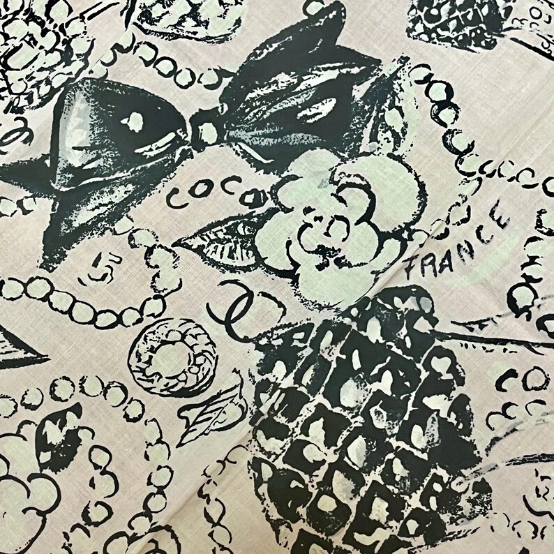 May include: A pink fabric with a repeating pattern of black and white illustrations. The design includes bows, camellias, pearls, and the word "France". The word "Coco" is also visible.