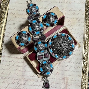 Op de afbeelding: Een antieke sieradenset met een armband, oorbellen en een broche. De zilverkleurige stukken hebben een filigraan ontwerp met turquoise en bordeauxrode accenten. De set wordt gepresenteerd in een vintage sieradendoosje.