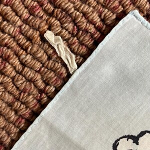 May include: A light blue handkerchief with a light blue border and a decorative design. The handkerchief is partially visible, resting on a textured brown and red surface. A small, cream-colored fabric piece is also present.